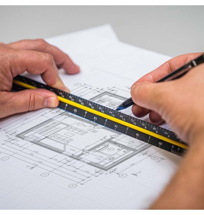 Hands using a GEN Aluminum Triangular Architect Scale and pen on floor plans with a grid layout and room illustrations. The scale's color-coded grooves enhance clarity, offering precision as the high-impact aluminum body glides over the 12-in scale of the architectural drafts.