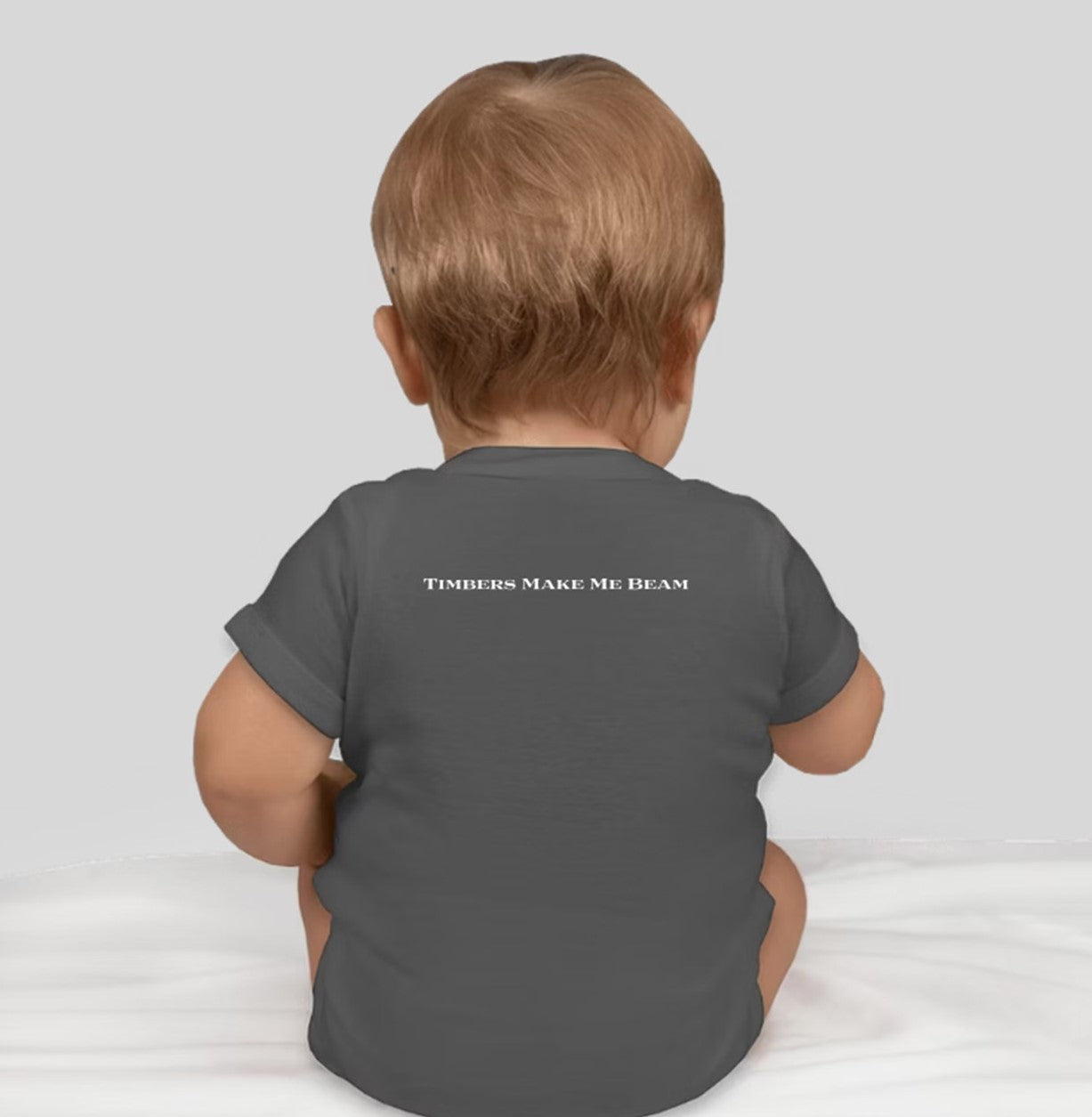 A baby sitting on a white surface, facing away, wearing a dark gray Custom Ink Shelter Onesie with text "TIMBERS MAKE ME BEAM" on the back.