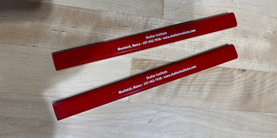 Two red pencils with "Shelter Institute Woolwich, Maine 207-442-7938 www.shelterinstitute.com" printed on their sides are laid out on a wooden surface—essentials in any seasoned timber framer's Deluxe Timber Framing Tool Kit: Best from Shelter Institute.