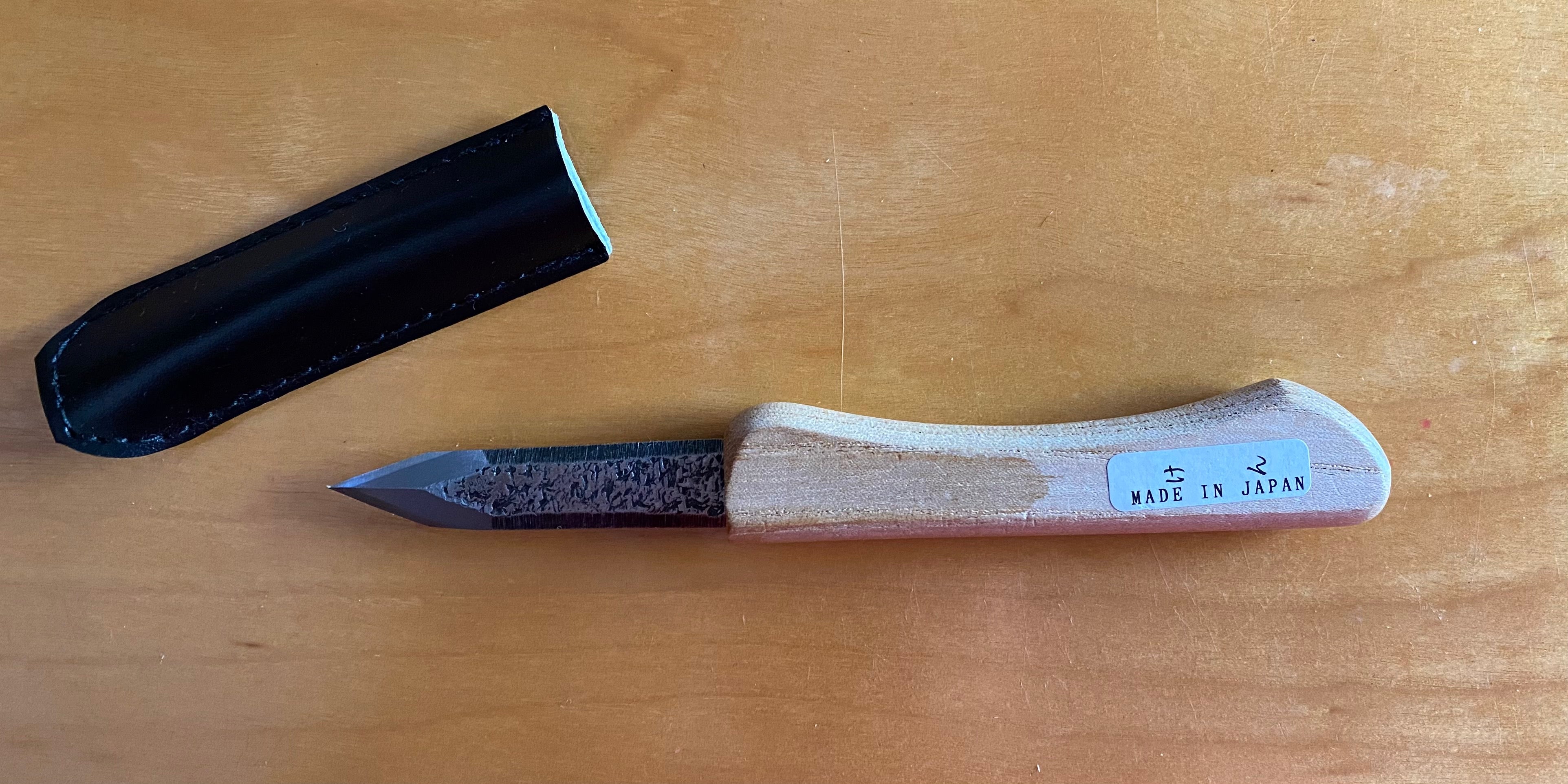 An Ikeuti Japanese Marking Knife from the brand MARU, featuring a laminated steel blade and wooden handle, rests on a wooden surface. This exquisite tool, proudly labeled "Made in Japan," includes a black sheath and showcases its double bevel precision for fine woodworking tasks.