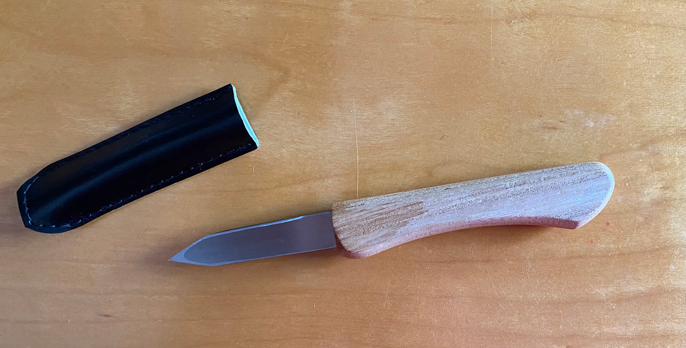 The Ikeuti Japanese Marking Knife by MARU, featuring a wooden handle and double bevel edge, rests beside its black sheath on a wooden surface.