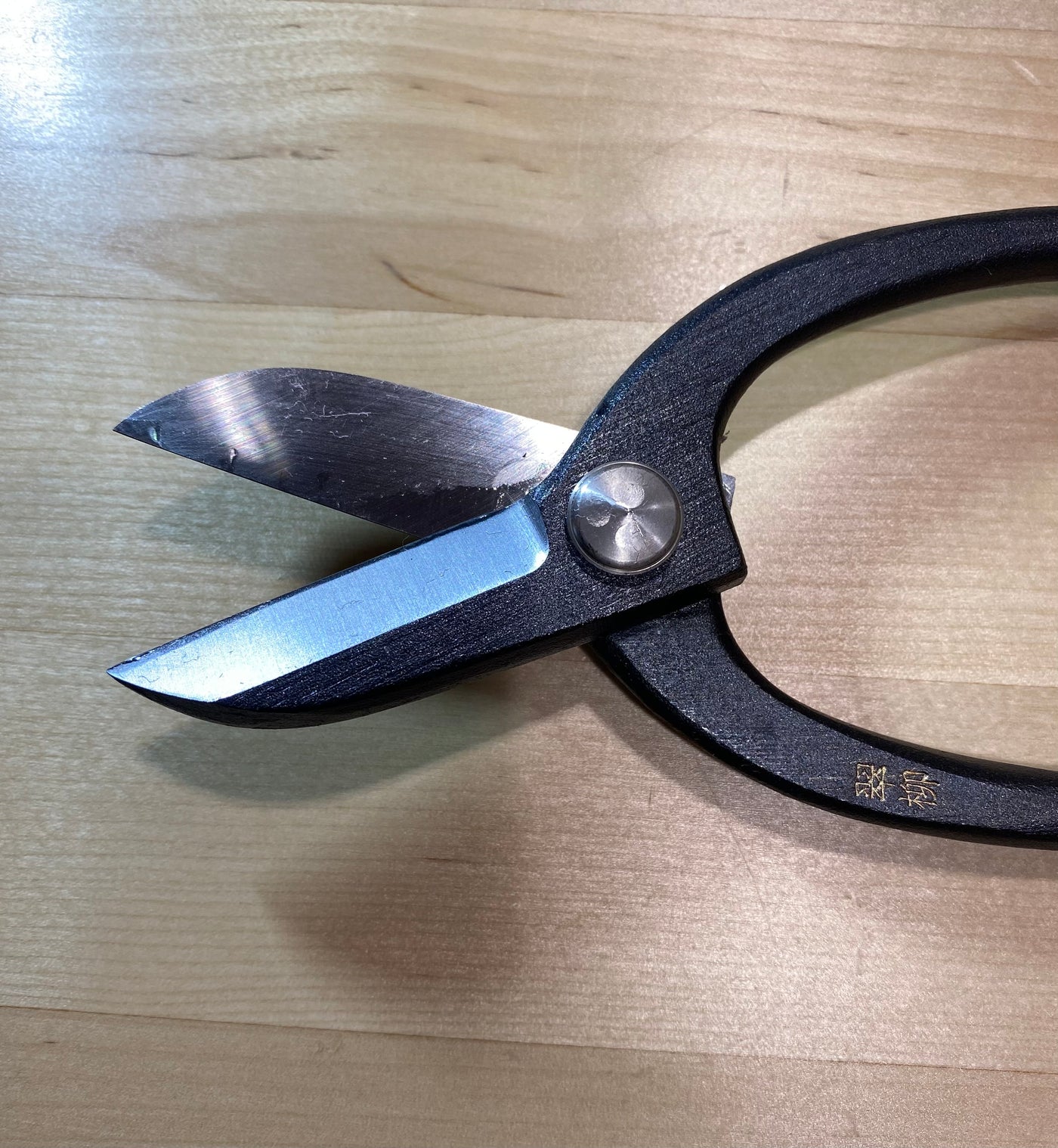 The MARU Ikebana Shears, with black handles and sharp carbon steel blades, are perfect for Japanese flower arranging and rest elegantly on a light wooden surface.