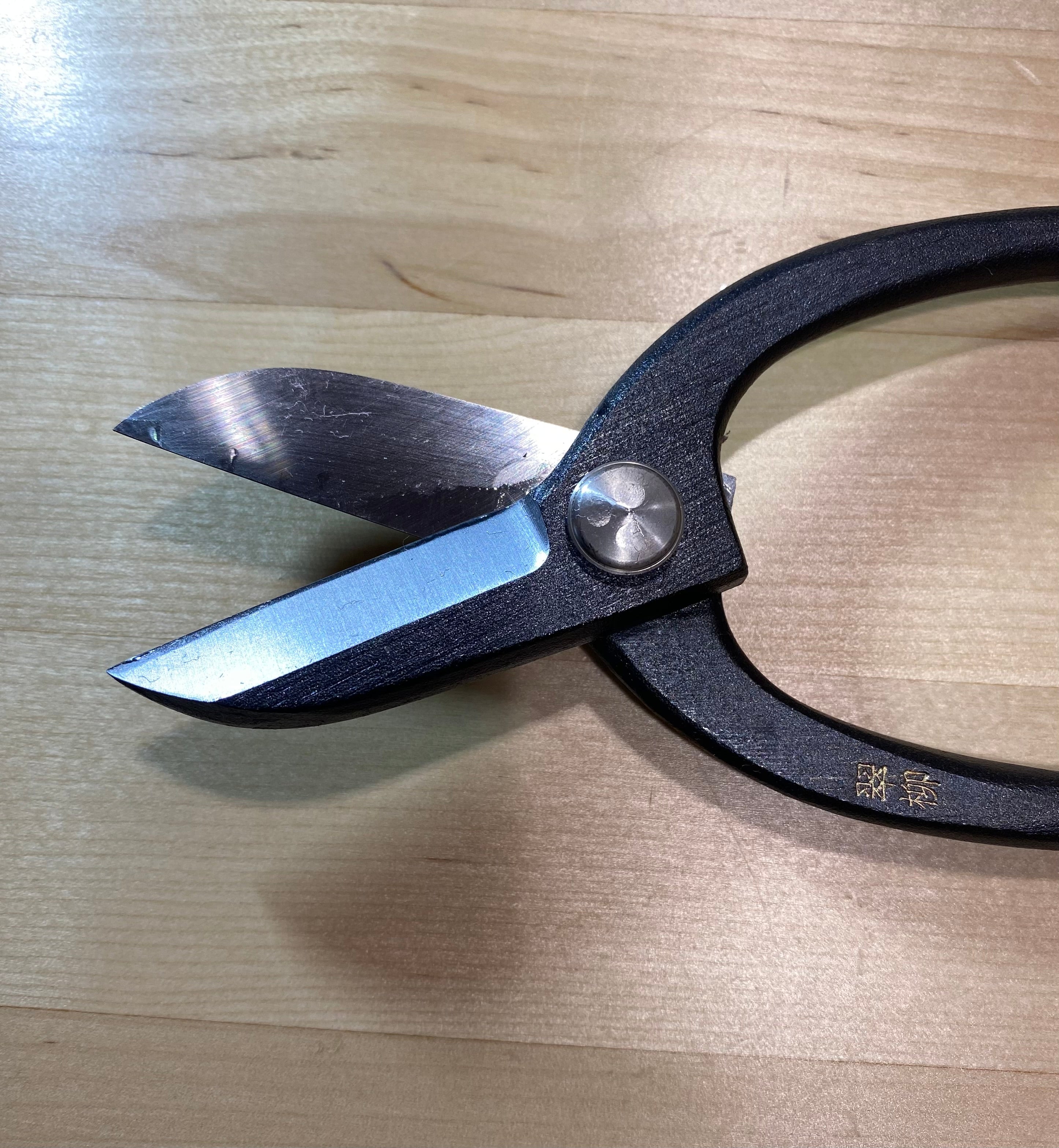 The MARU Ikebana Shears, with black handles and sharp carbon steel blades, are perfect for Japanese flower arranging and rest elegantly on a light wooden surface.