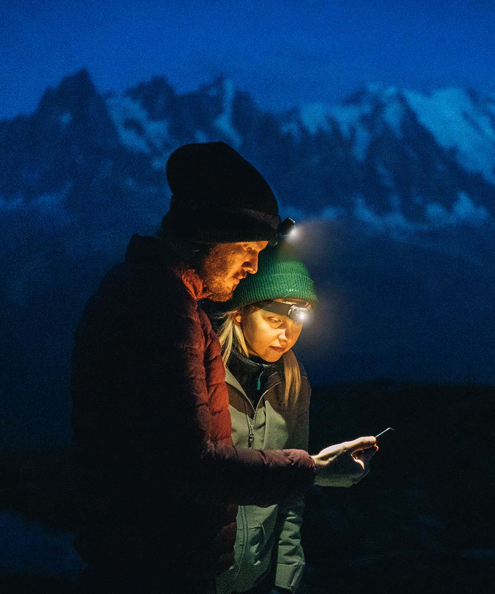 Two people, each wearing a NEB Nebo Mycro Headlamp with adjustable head straps, stand together at night, looking at a device. Snow-covered mountains are visible in the background.